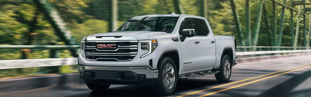 The image features a GMC Sierra 1500 driving across a bridge surrounded by lush greenery. The truck is captured in motion, showcasing its sleek, modern design.