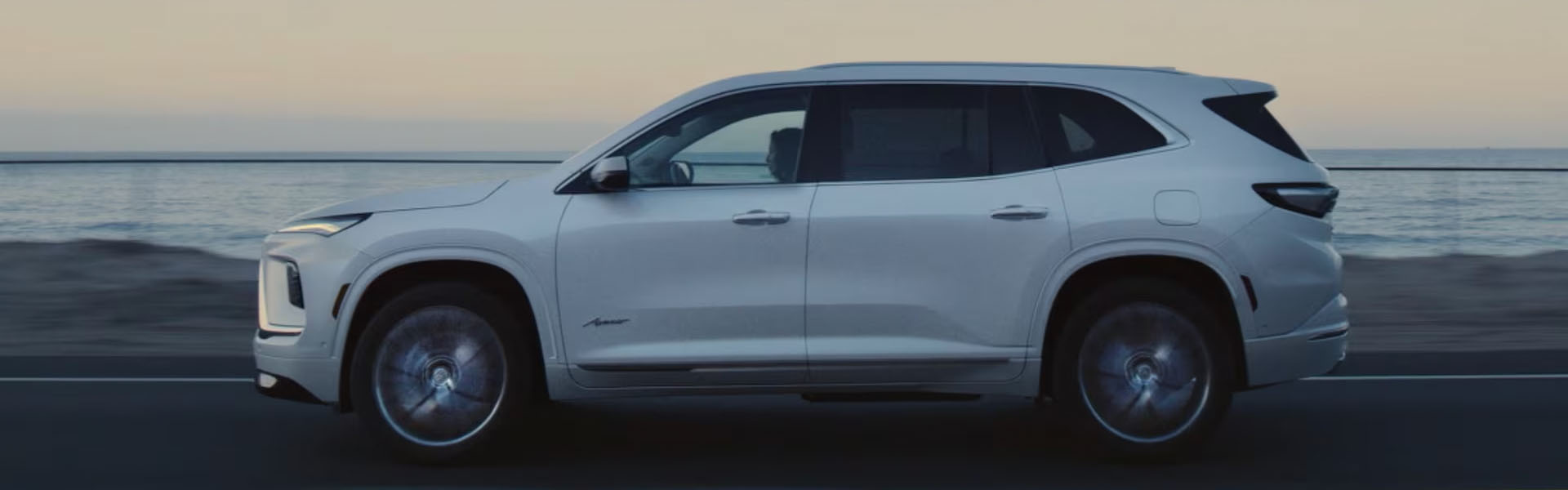 2026 Buick Enclave SUV driving along a coastal road at dusk, shown in side profile with the ocean in the background.